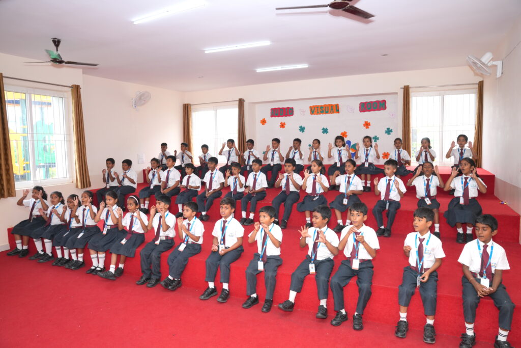 Aadya Academy Budigere students seated on tiered carpeted steps practicing hand gestures in an audio-visual room