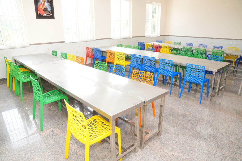 Clean and spacious student dining hall at Aadya Academy Budigere featuring stainless steel long tables and vibrant colorful chairs